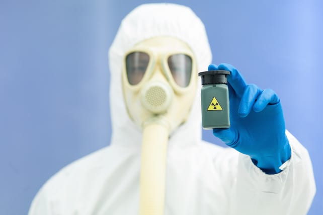 person in bio hazard suit and gas mask holding a radioactive isotope sample in a small green plastic jar